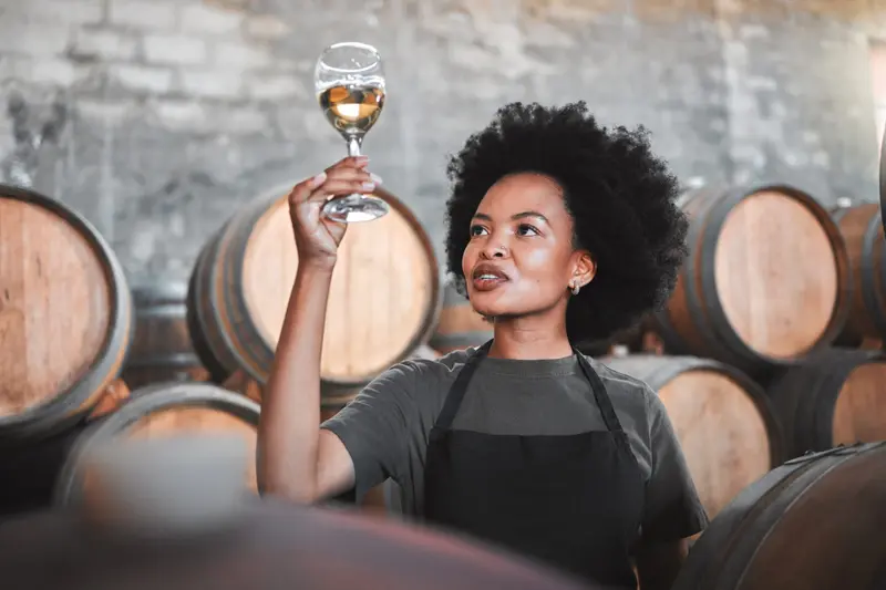 A woman in an apron holding a glass of white wine between barrels in a cellar