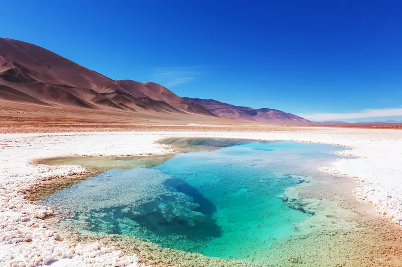 A turquoise pool on the salt flat, with dry hills behind it