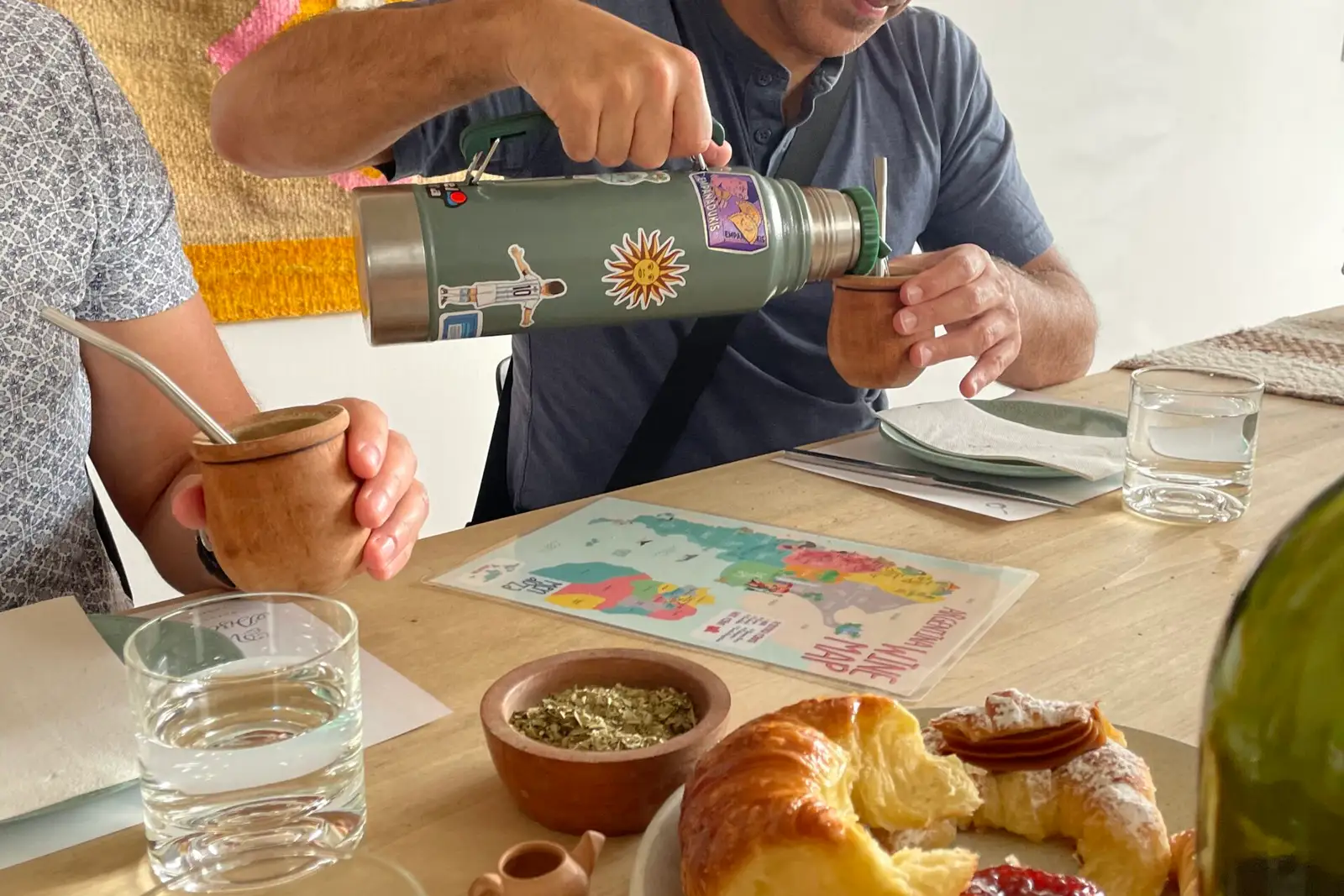 Pouring from a thermos flask into a mate, at a table laid with pastries
