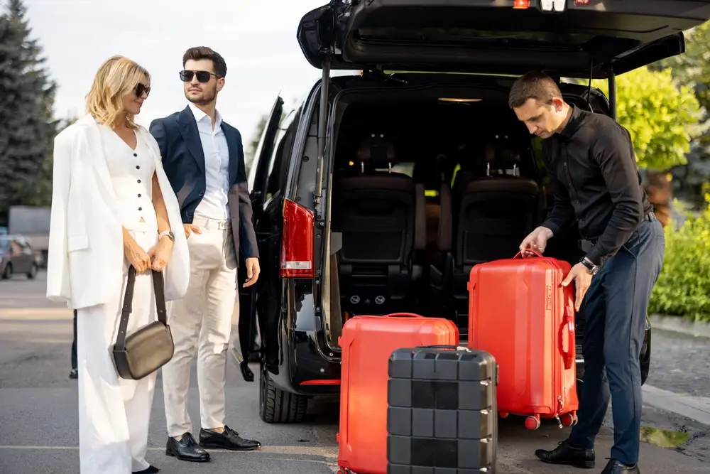 A driver loading suitcases into the back of a van, with the passengers beside it